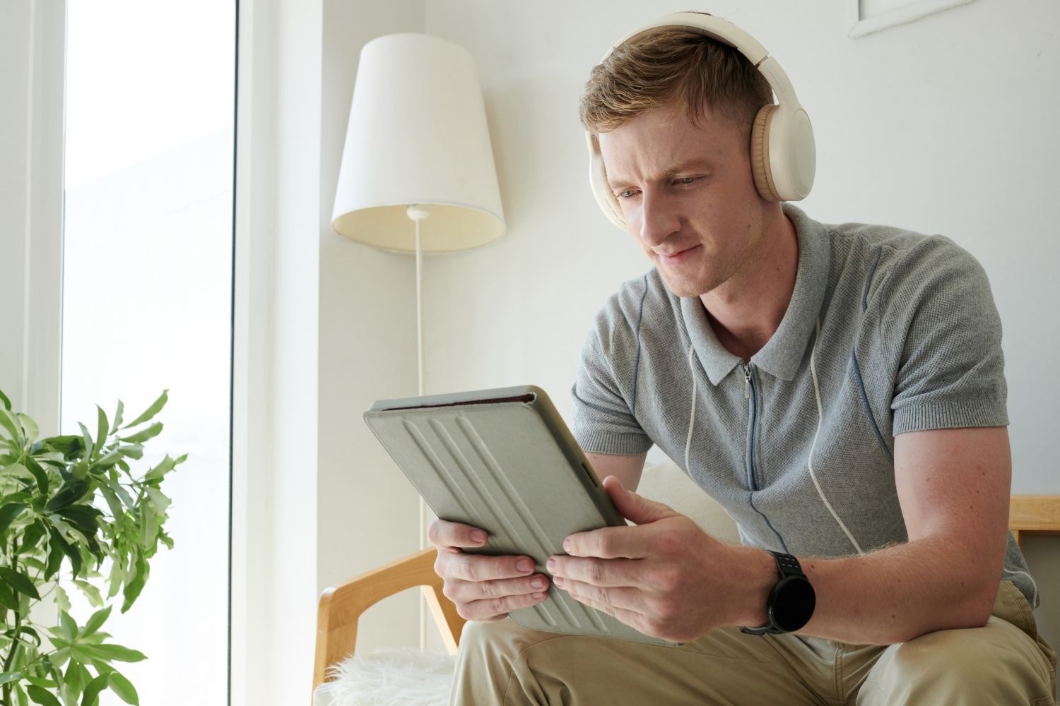 En Maa luegt konzentriert uf sis Tablet mit-re IPTV Player App, während er Chopfhörer trait und uf-ere Holzcouch sitzt, naben-ere grossen Pflanze und emne Fänschter.